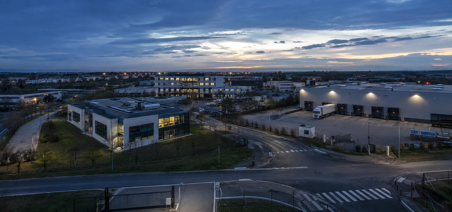 Vue d'un pôle d'activités français à l'heure bleue où se lisent bureaux, logistique, routes et accès : la diversité des environnements couverts par PREGERIS.